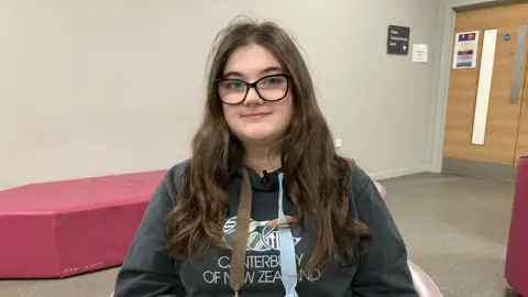 Leanne Rinne/BBC A young girl sits on a chair looking into the camera smiling. She is wearing large blacks glasses and is wearing a dark hooded jumper. She has long brown hair. 