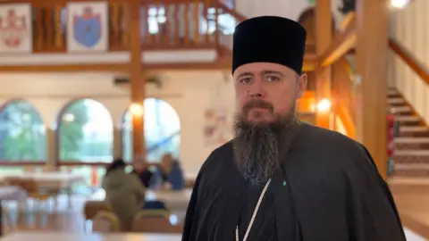 A man wearing black religious attire which includes a black cloak, pillar shape hat and a chain. He has a beard and moustache and is looking at the camera. Behind him are people, wooden stairs and paintings which are out of focus. 