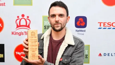 A man with a short brown beard and short brown hair, wearing a wool lined grey denim jacket, looks towards the camera while holding a wooden King's Trust award