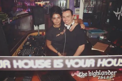 Hannah Laing A girl in a black top with black hair has her arm around a man wearing a black top and white headphones. They both stand in a DJ booth in a bar.