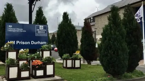 BBC View of a sign for HMP Durham with flower boxes in front of it, and to the side a partial view of the brick structure with small trees in front of it.
