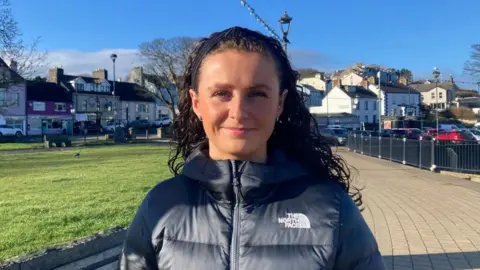 Maria has black curly hair and is wearing a black North Face coat. She is looking directly at the camera and is standing in front of a car park.