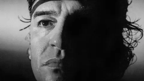 Adam Docker A close up photo of Diego Maradona, half his face is obscured by shadow. 