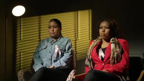Elise Skillen and Savannah Holm-Aderemi are sitting beside each other, in a panelled room, with a yellow light shining against the wall. Skillen is wearing a denim jacket and Holm-Aderemi has a red jacket and a patterned scarf around her neck. Both are wearing pink ribbons in tribute to Fiona Holm