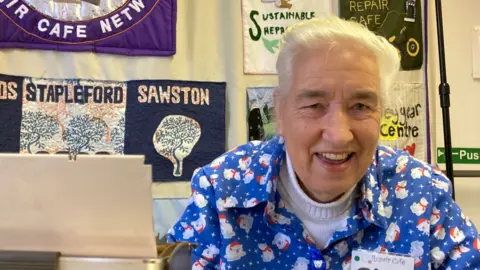 Gina wears a blue Christmas shirt and is concentrating on pushing a needle and thread through material - on the left is the arm of a sewing machine