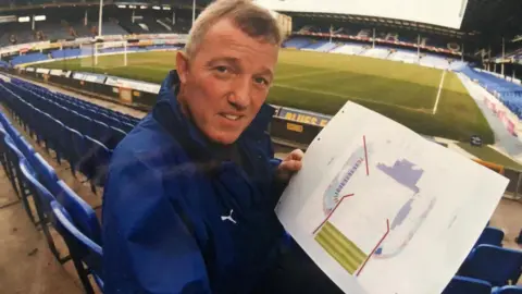 Everton FC Bob Lennon, pictured when aged in his 40s or early 50s, sits in an empty football stadium on a blue plastic seat holding a diagram showing the pitch. He is wearing a blue coat. 