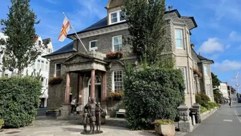 BBC The exterior of the States Offices on a blue sky day. There are sculptures of a man, woman and two children in the square in front of the building. There is a tree, bush and granite planter of flowers either side of the entrance. The entrance has a portico with columns which has the Guernsey flag flying from it. The building has three storeys and stands next to a road.