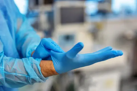 A stock image of a hand pulling on a light blue latex glove, the arm of the hand is dressed in medical scrubs