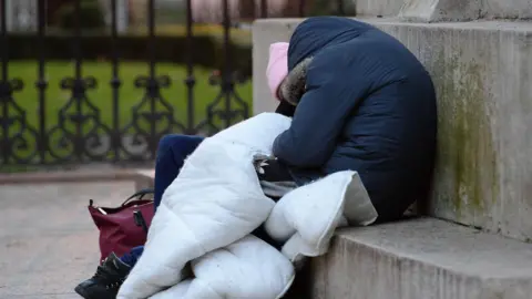 PA Media Two people sat on a concrete seat. Both are hunched over in thick jackets with their hoods up. Their legs are covered with a duvet to keep them warm. 