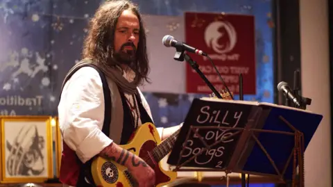 Graham Stubbs/Asylum Artist Quarter CIC A man playing a guitar in front of a microphone. He has long hair, a beard and is wearing a white shirt and waistcoat. In front of him is a book which says 'Silly Love Songs' on it. 
