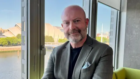 A man in a grey suit stands in an office beside the river Clyde