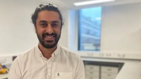 Ali Abdi smiles at the camera. He wearing a white shirt and is sat in a laboratory. 