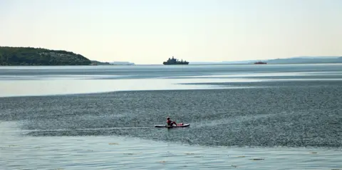 Helen Sadie A person in a canoe in the foreground with a large navy ship in the far distance 