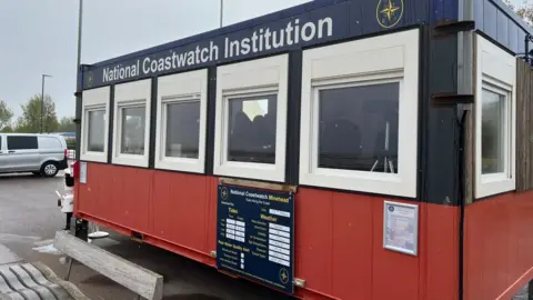 The outside of the blue and red National Coastwatch Institution cabin. A few cars can be seen in the background. The image was taken on an overcast day.