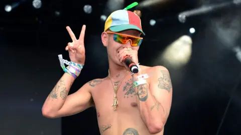 Slowthai performing at a festival. He is wearing a multicoloured hat and sunglasses and is singing into a microphone. He is shirtless, showing several tattoos on his torso and arms. He is making a peace sign with his right hand.