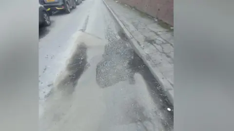 Matt Hunton A rough patch of concrete and aggregate sits in the middle of a residential road, contrasting with the darker tarmac around it. Cars are parked along the left side of the street, with a brick wall and pavement running along the right. The road surface is wet and shows signs of general wear and cracking.
