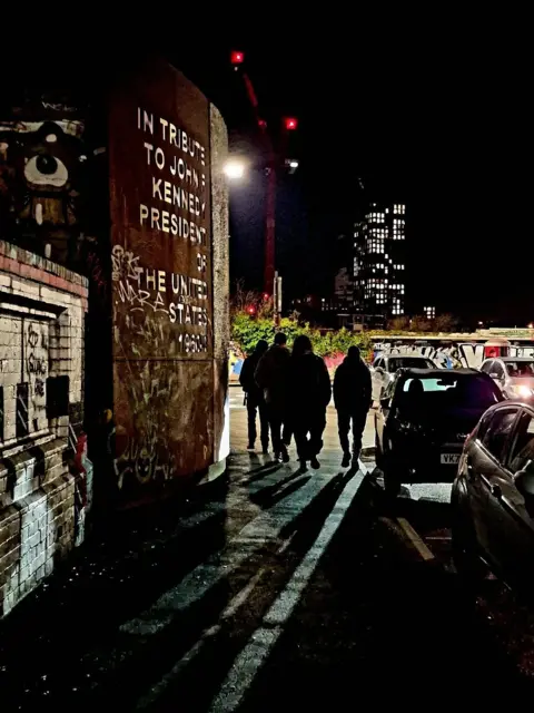 @photosbyvaleb1 Four people walk away from us along a pavement with long shadows behind them, it is nighttime. To the left of them is a red-brown wall with stenciled writing on it while to the right are several cars. In the distance, the lights of a tower block can be seen.
