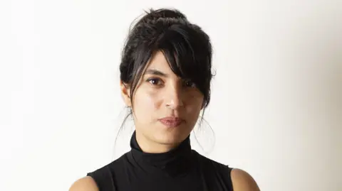Yasmin poses in sleeveless black high-neck top. She has long black hair worn up and a fringe and dark eyes.