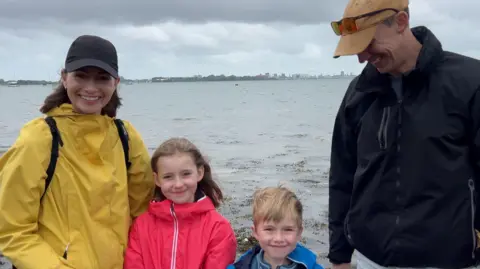 Image show a mum, dad, and two children smiling at the camera. Bheind them, you can see the water and Portsmouth. From left to right, the mum is wearing a yellow jacket, a baseball cap and has brown shoulder length hair. The young girl is stood next to her mum. She is wearing a pink waterproof jacket. Her hair is blowing in the wind and she is smiling at the camera. Her brother is next to her and is wearing a blue waterproof jacket and a blue striped quaterzip. He has blond hair. Next to the young boy, is their dad. He is wearing a mustard coloured that with sunglasses on top and a black waterproof jacket. He is smiling and looking down at his son. 