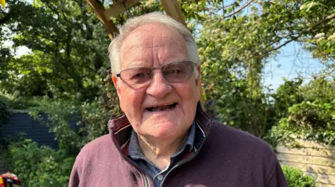 Dafydd Iwan looks straight to camera, smiling with his mouth open. He has white cropped hair and tinted glasses. He wears a maroon zip jumper over a grey shirt.