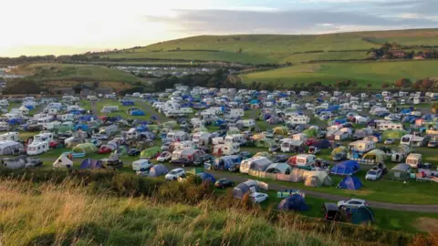 Aerial photo of a field with hundreds of tents pitched in it with a grassy hill in the background