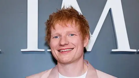 Ed Sheeran smiles for photographers during an event. He has red hair and wears a salmon pink suit jacket with a white T-shirt underneath.