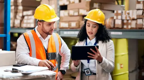 Getty Images A man and a woman are talking inside a factory. They both have yellow safety helmets on and the woman is holding a small computer. The man is also wearing a fluorescent jacket.