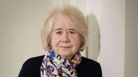 Kelvin Boyes / PressEye Anne Cadwallader is an older woman with very light blonde hair. She is standing in front of a white wall, wearing a black top with a multi-coloured silk-type scarf draped around her neck. 
