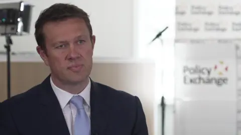 David wears a dark suit and a blue tie. He is sitting in a conference room, with the Policy Exchange logo behind him, 