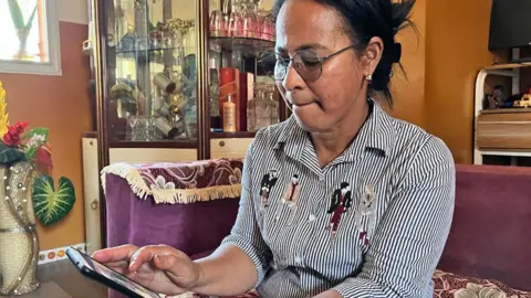 Sammy Awami/BBC Razanasoa Edmondine, sucking her lips, wears a black-and-white striped long-sleeved shirt and looks at recent photos of her grandson at her home near Antananarivo.