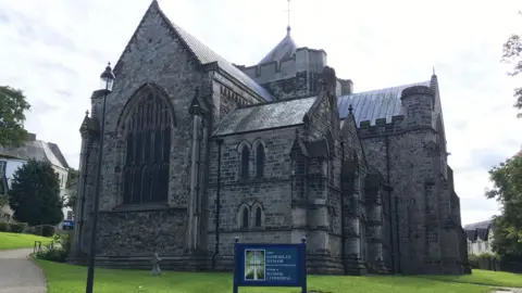 A picture of the front of the large Bangor Cathedral on a bright day with a blue sign at the forefront of the picture