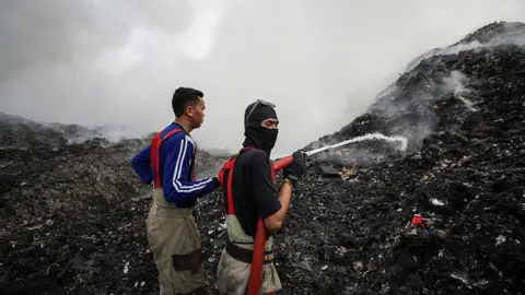 Two men, one wearing a black balaclava, spray water from a hose onto a black smouldering hillside