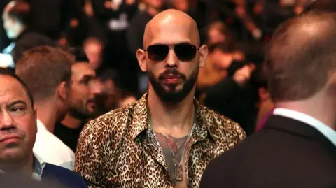 Andrew Tate wearing dark sunglasses and a button-down animal print shirt and a dogtag necklace