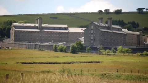 A wide shot of Dartmoor Prison. It is a row of dark grey buildings surrounded by green fields.