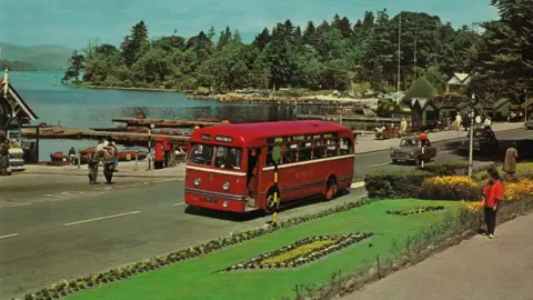 Postcard dating from the 1960s showing - in highly saturated colour - a red single decker bus on a road next to the shore of a lake. A number of pedestrians are strolling on the pavement, next to a floral display in a grassed area and there are rowing boats tied to jetties on the lake.