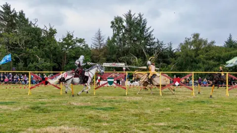 HES Two knights charge at one another with a side-on view and trees in the background