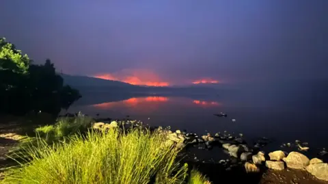 Malcolm Smith It is dark and flames glow orange on the horizon. The flames are reflected in the mirror-like surface of Loch Morlich.
