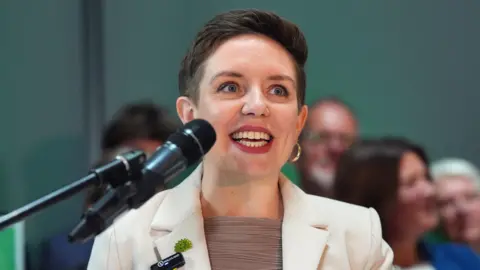 PA Media Carla Denyer standing behind a microphone ready to speak. She is wearing a cream jacket and brown top, with red lipstick and gold hoop earrings.