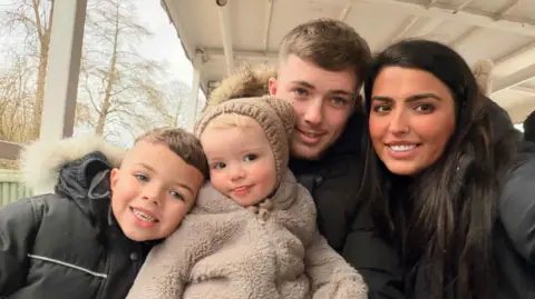 Family photo Family photo of two adults and two small children. The boy on the left is wearing a black coat with a furry hood. He's next to a little girl in a furry, light brown coat. She's wearing a matching hat. A man is next to them. He has light brown hair. On the right is a woman with long dark hair wearing a black coat. They're all smiling. 