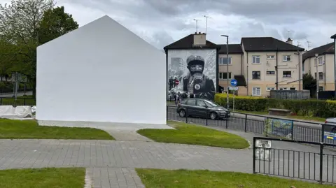 Free Derry Corner is painted white. A car is visible in the background as well as a number of houses.