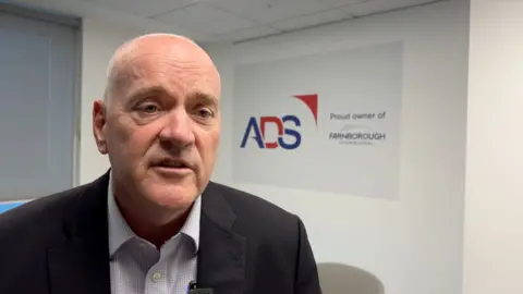 A man in a dark jacket and open knecked shirt sits inside an office with the logo of ADS, the industry trade body behind the airshow, on the wall behind him.