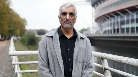 BBC Sarju is wearing a black shirt and a grey over-shirt. He is looking at the camera with a neutral expression. Behind him is the River Taff and the Principality Stadium.