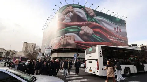 AFP via Getty Images People walk past a large patriotic banner depicting the Iranian flag on Enghelab Square in Tehran on January 14, 2026