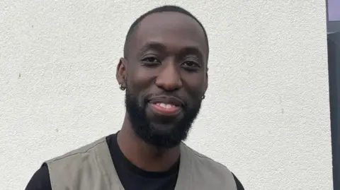 A man in a black t shirt and grey waistcoat stands in front of a grey wall. he has short black hair and beard and is smiling