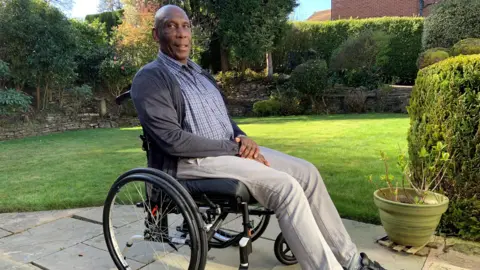 Uriah Rennie sits in a wheelchair. He is sitting sideways and looking into the camera. He is wearing a shirt, cardigan and trousers