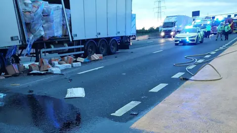 National Highways  Emergency service vehicles are at the scene, the large white lorry to the left has the side broken open and boxes are on the floor and spilling from the side