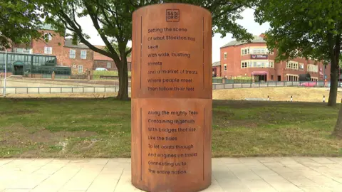 A rust coloured steel tube stands in the middle of a pathway. Below a symbol for the Stockton and Darlington Railway, the following words have been engraved: "Setting the scene of what Stockton has been. With wide, bustling streets and a market of treats. Where people meet then follow their feet along the might Tees. Containing ingenuity, with bridges that rise like the tides to let boats through. And engines on track, connecting us to the entire nation.