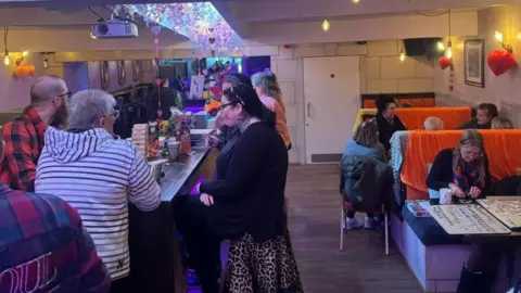 A cafe with booths along the right wall is full of people chatting or doing jigsaws.