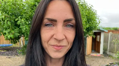 A woman with long brown hair stands in a garden. She is smiling at the camera.