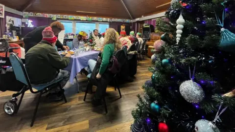 People sitting at tables enjoying a Christmas lunch. They are wearing Christmas hats and there is a Christmas tree in the foreground.